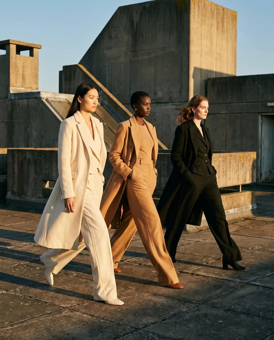 Drie modellen in crème, camel en zwart lopen over een betonnen dak in laag zonlicht